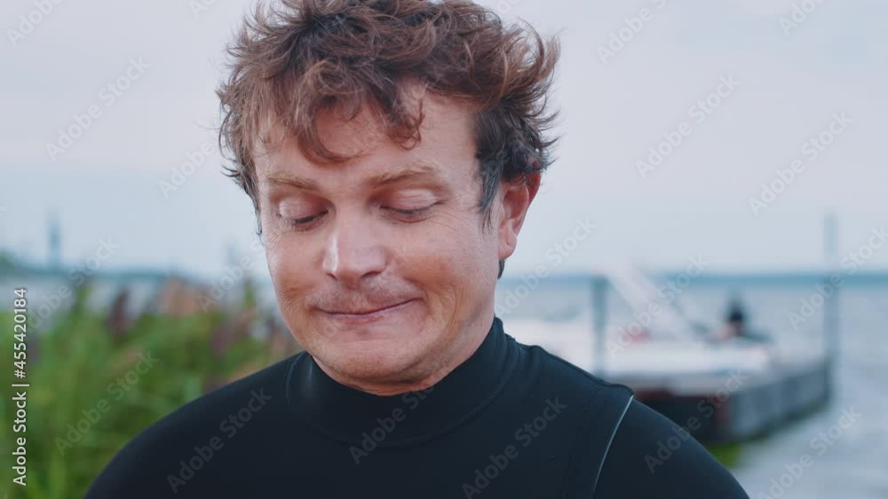 Wake surfer interview. Portrait of the talking man in black wetsuit standing outdoor with lake and boat on the background