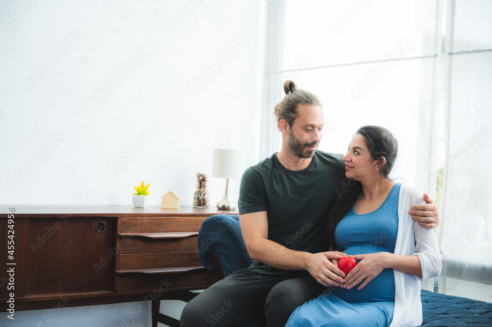 Beautiful young pregnant woman sitting on bed with loving and caring husband trying to connect with baby by touching and feeling stomach at home Beautiful young pregnant woman sitting on bed with loving and caring husband trying to connect with baby by touching and feeling stomach at home