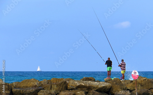  Two adult men are fishing with fishing rods in the sea from the coastal rocks. A sailing yacht is visible on the horizon. 