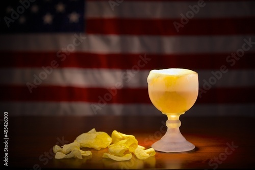 Photography Ice cold schooner glass with beer in front of an American flag with some potato chips on the side