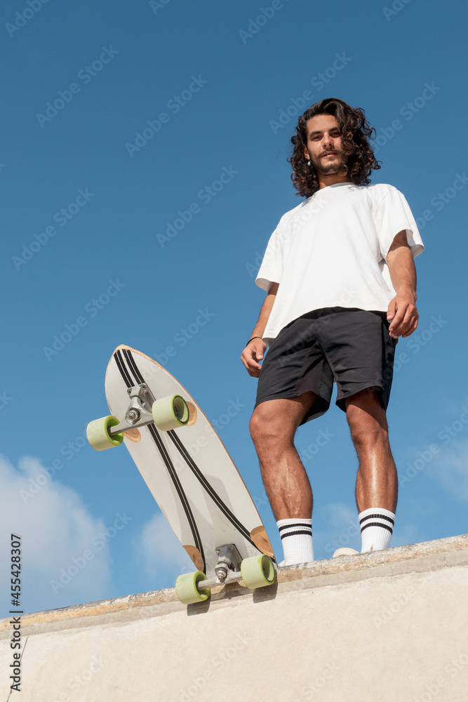 Young handsome skater standing at the top of the skate ramp. He has long  black hair and he is wearing summer clothes. Photos | Adobe Stock