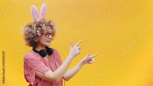 Attractive young girl wearing cute bunny ears and spectacles in a joyful mood. Headphones around her neck. Girl smiling and pointing at something. photo