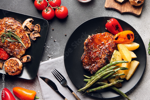 Plates with tasty pork steaks and grilled vegetables on grey background