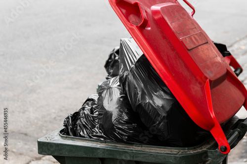Black plastic bags over flowing modern trash bin with red lid.