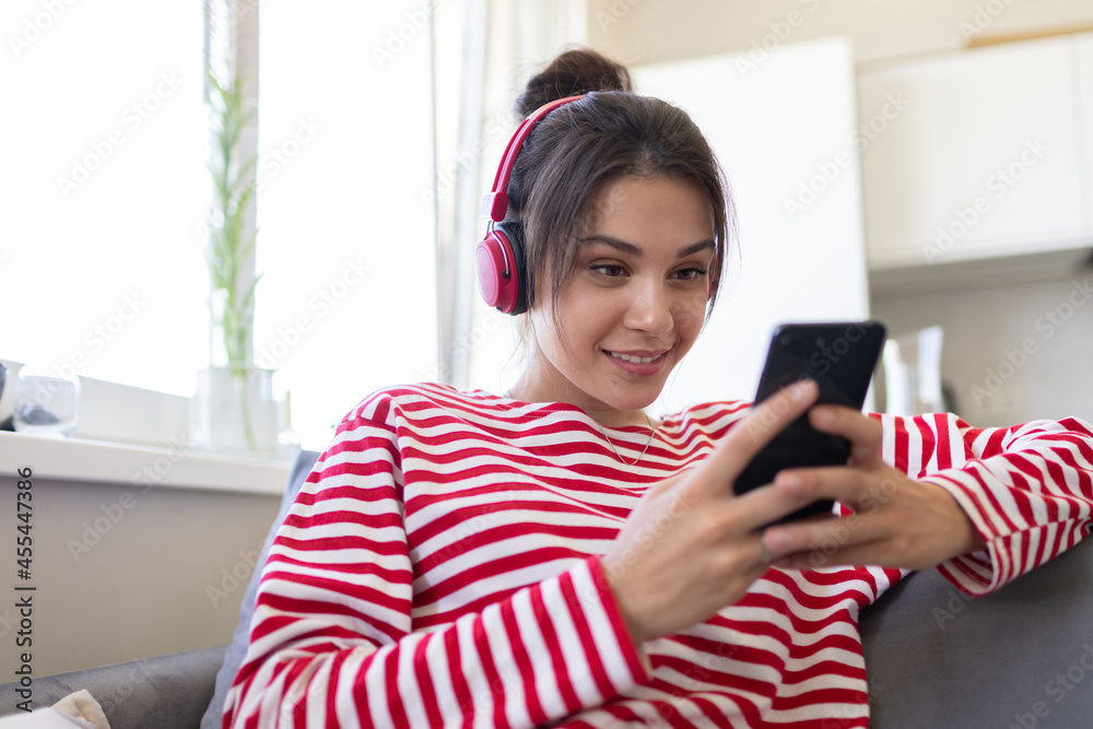 Girl in wireless headphone with phone using mobile player app listening online music. Female learning language, watching movie at home, communicating in social networks, domestic weekend pastime Girl in wireless headphone with phone using mobile player app listening online music. Female learning language, watching movie at home, communicating in social networks, domestic weekend pastime