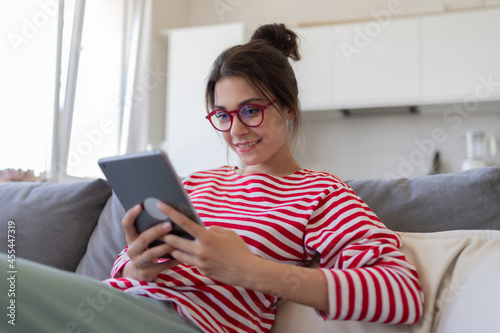 Modern happy girl with tablet computer browsing internet. Smiling female in eyeglasses checking email, shopping or learning online sitting on sofa at home.