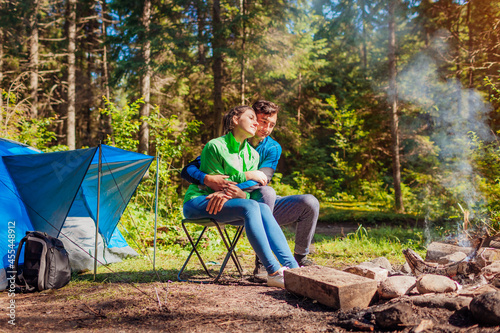 Young couple of travelers enjoy campfire in summer forest. Tourists sitting by tent hugging. Camping in woods