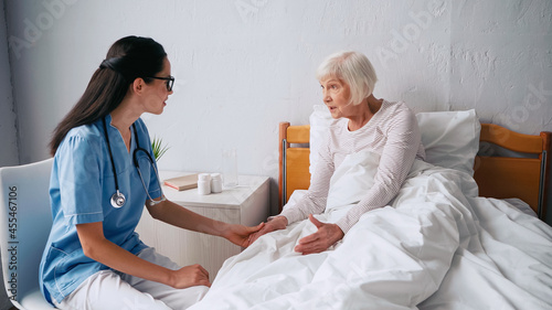 geriatric nurse in eyeglasses talking with aged woman in bed