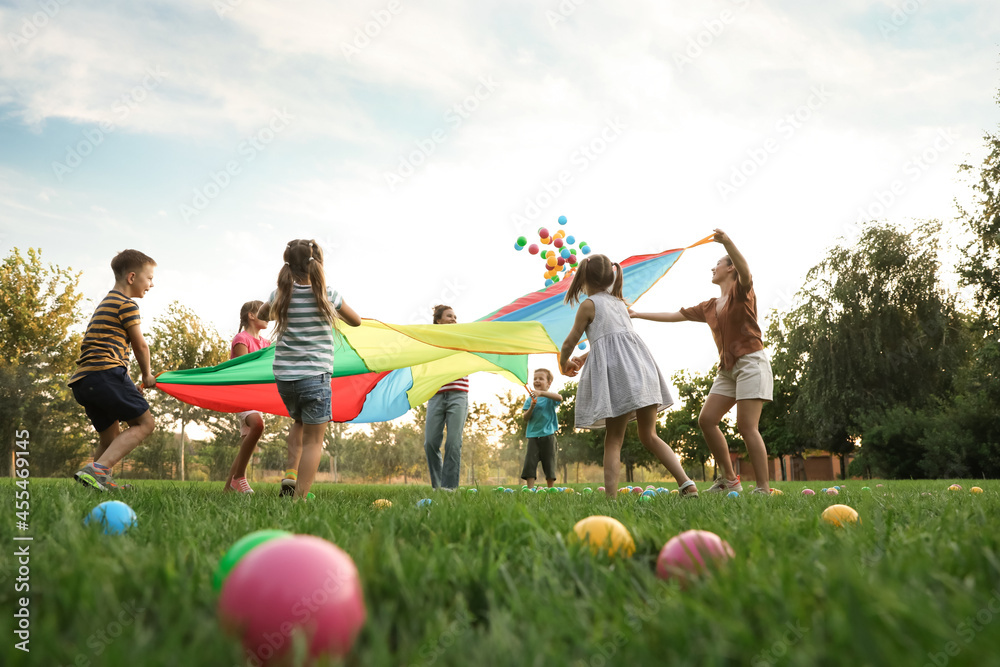 Group of children and teachers playing with rainbow playground parachute on green grass, low angle view. Summer camp activity - obrazy, fototapety, plakaty 
