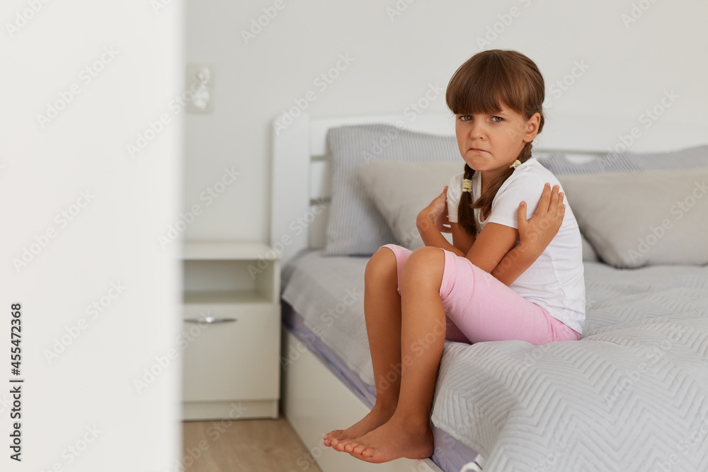 Portrait of charming offended sad female kid wearing white t shirt and pink short, looking at camera, crying, feeling sorrow, posing on bed in light room alone. Portrait of charming offended sad female kid wearing white t shirt and pink short, looking at camera, crying, feeling sorrow, posing on bed in light room alone.