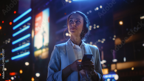 Beautiful Young Woman Using Smartphone Standing on the Night City Street Full of Neon Light. Portrait of Gorgeous Smiling Female Using Mobile Phone. photo
