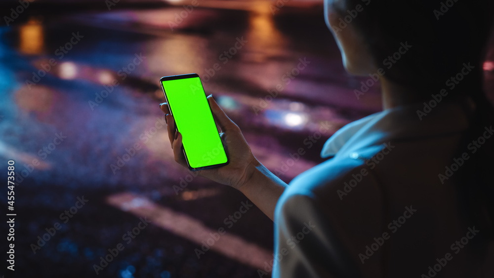 Obraz premium Beautiful Woman using Chroma Key Smartphone while Walking Through Night City Street Full of Neon Light. Female Using Green Screen Mobile Phone. Over the Shoulder Shot
