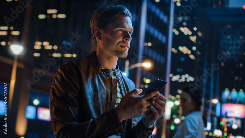 Portrait of Handsome Young Man Using Smartphone Standing in the Night City Street Full of Neon Lights. Smiling Stylish Blonde Male Using Mobile Phone for Social Media Posting. Dutch Angle Shot. © Gorodenkoff