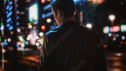 Following Shot of Handsome Man Using Smartphone Walking in a Modern City Street with Neon Lights at Night. Attractive Male Walking Through Urban Area using Mobile Phone.