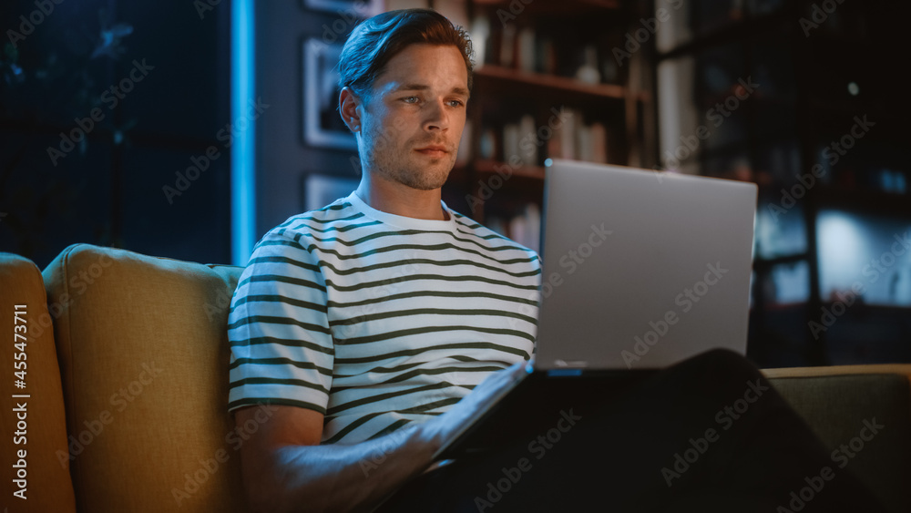 Man Using Laptop at Home Office, Does Remote Work in the Evening. Handsome Male Sitting on Sofa Works On Computer, Brainstorms Creative Project, does Online Shopping, Browsing Internet. Man Using Laptop at Home Office, Does Remote Work in the Evening. Handsome Male Sitting on Sofa Works On Computer, Brainstorms Creative Project, does Online Shopping, Browsing Internet.