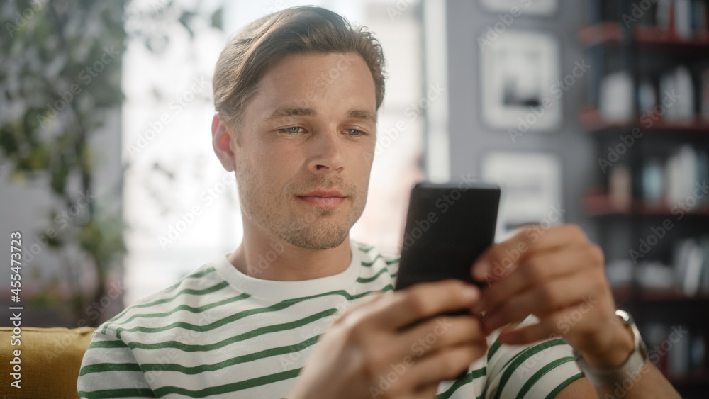 Sunny Day: Happy Handsome White Man Using Smartphone in Cozy Living Room at Home Sitting on a Sofa. Doing Online Shopping, Browsing the Internet and Checking Videos on Social Media. Close-up Shot Sunny Day: Happy Handsome White Man Using Smartphone in Cozy Living Room at Home Sitting on a Sofa. Doing Online Shopping, Browsing the Internet and Checking Videos on Social Media. Close-up Shot