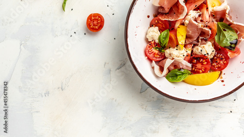 White bowl of fresh and healthy Mediterranean salad with tomatoes, cream cheese balls, prosciutto jamon and basil leaves. Delicious balanced food concept. Long banner format. top view