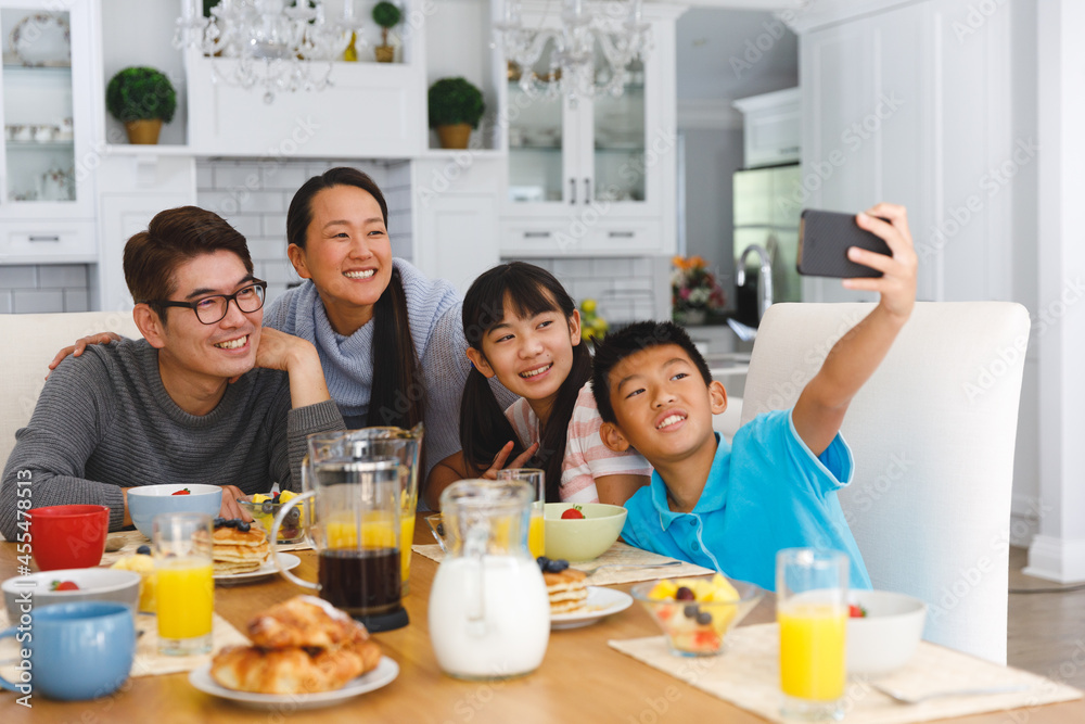 Smiling asian parents with son and daughter sitting at breakfast table taking selfie