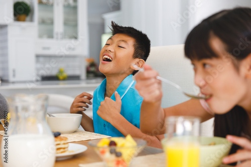 Laughing asian brother sitting at breakfast table with sister eating