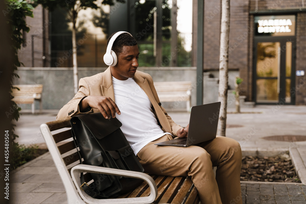 Young black man in headphones working with laptop while sitting on bench Young black man in headphones working with laptop while sitting on bench