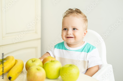 cute baby boy sitting at the table in child chair eating apple on white kitchen.