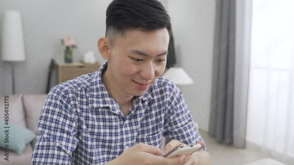 close up Taiwanese guy in plaid shirt is smiling while sending text message to his friend on the cell phone at home. close up Taiwanese guy in plaid shirt is smiling while sending text message to his friend on the cell phone at home.
