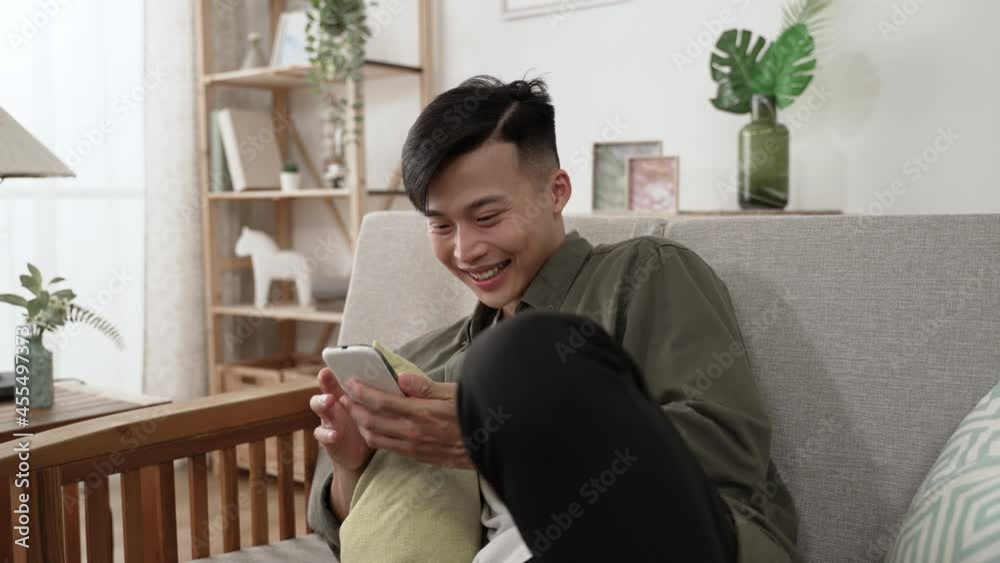 carefree asian young man throwing head back and laughing loudly while watching a funny video on his mobile phone on comfortable sofa in the living room at home. carefree asian young man throwing head back and laughing loudly while watching a funny video on his mobile phone on comfortable sofa in the living room at home.