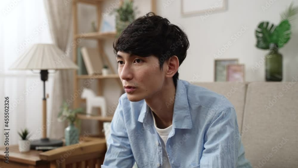 depressed asian young man sitting on the living room sofa in silence and showing no expression on face while staying at home alone during self isolation.