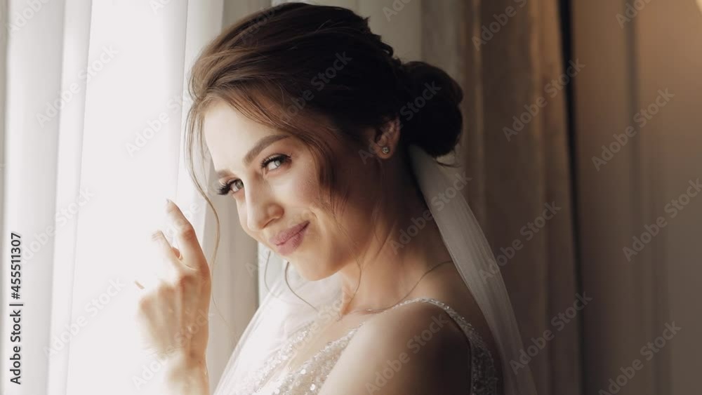 Bride in white dress staying near window, waiting for groom. Wedding morning preparations. Bride with fashion hairstyle and makeup in hotel or home apartment. Beautiful, sexy woman in gown and veil Bride in white dress staying near window, waiting for groom. Wedding morning preparations. Bride with fashion hairstyle and makeup in hotel or home apartment. Beautiful, sexy woman in gown and veil