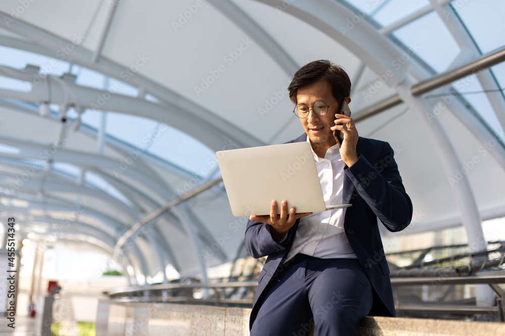 Asian confidently business man in glasses and suit working on laptop and mobole phone outdoor in city.