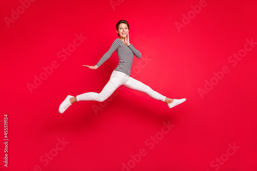 Photo of cute excited young lady wear striped clothes smiling jumping high arm cheek isolated red color background