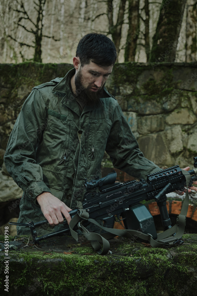 Portrait of airsoft player in professional equipment loads a gun with bullets in the forest. Soldier with weapons at war