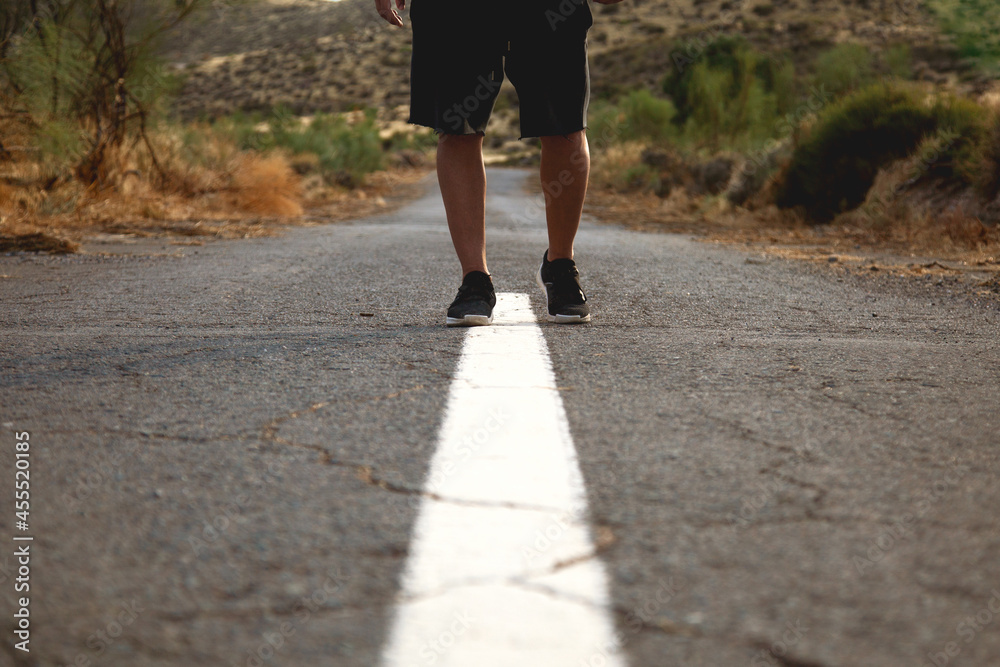 Hombre andando sobre línea continua en una carretera de asfalto desértica con fondo de montañas y plantas. Clima de verano. Hombre deportista en carretera. 