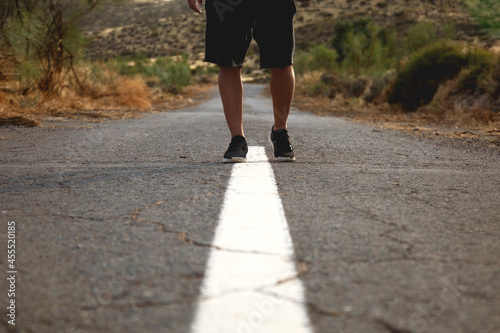 Hombre andando sobre línea continua en una carretera de asfalto desértica con fondo de montañas y plantas. Clima de verano. Hombre deportista en carretera.  photo