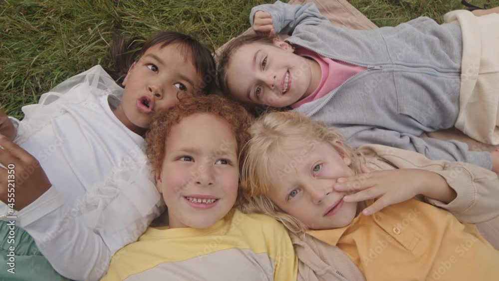 Top view POV of four happy multiethnic grade schoolers lying heads together on lawn, smiling and looking on camera Top view POV of four happy multiethnic grade schoolers lying heads together on lawn, smiling and looking on camera