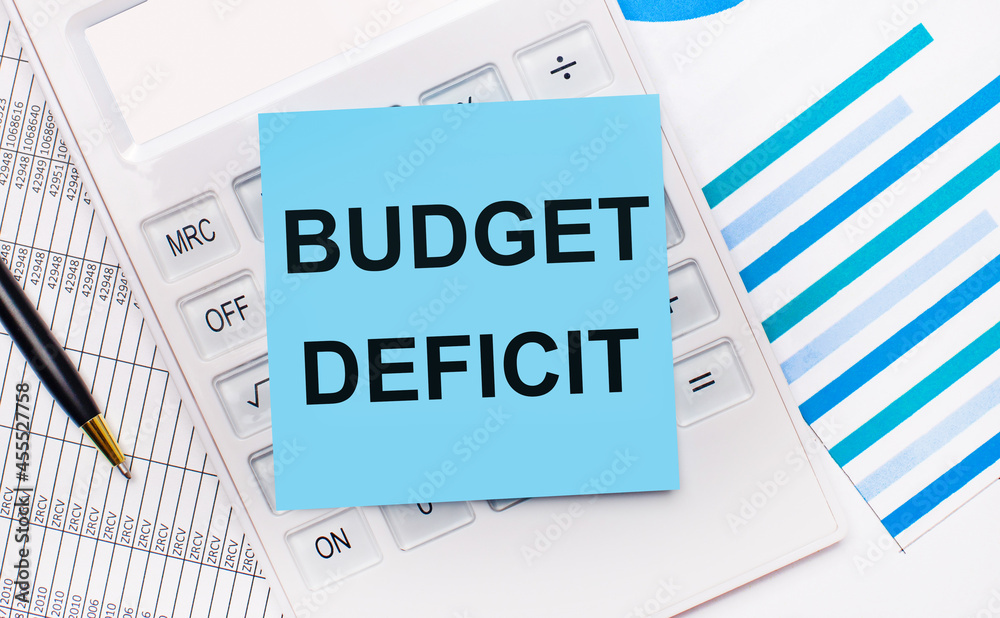 On the desktop is a white calculator with a blue sticker with the text BUDGET DEFICIT, a pen and blue reports. Business concept On the desktop is a white calculator with a blue sticker with the text BUDGET DEFICIT, a pen and blue reports. Business concept
