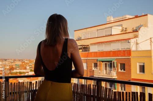 Mujer disfrutando de su terraza mirando a través de ella con edificios al fondo. Madrid desde el cielo. Mujer de espaldas. Chica con camiseta tirantes negra y falda amarilla