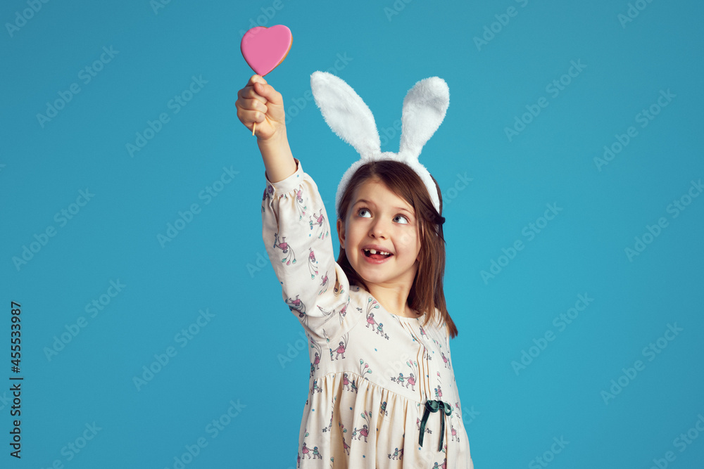 Cheerful girl smiling and holding tasty cookie in shape of heart, wearing bunny ears and cute dress, isolated over bright blue color background