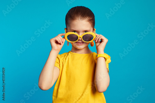 Lovely little girl in yellow shirt, looking at camera while adjusting sunglasses, isolated over blue background