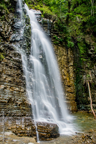 A high and powerful waterfall deep in the forest  surrounded by grass  rocks  trees. The waterfall gives rise to the river. Very beautiful wallpaper with a landscape and beautiful nature.