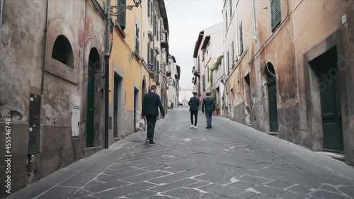 Back view of Italian citizens walking along the old narrow medieval street with characteristic building exterior, buildings with ruined facades of various architectural epoch. Antique Italian villages