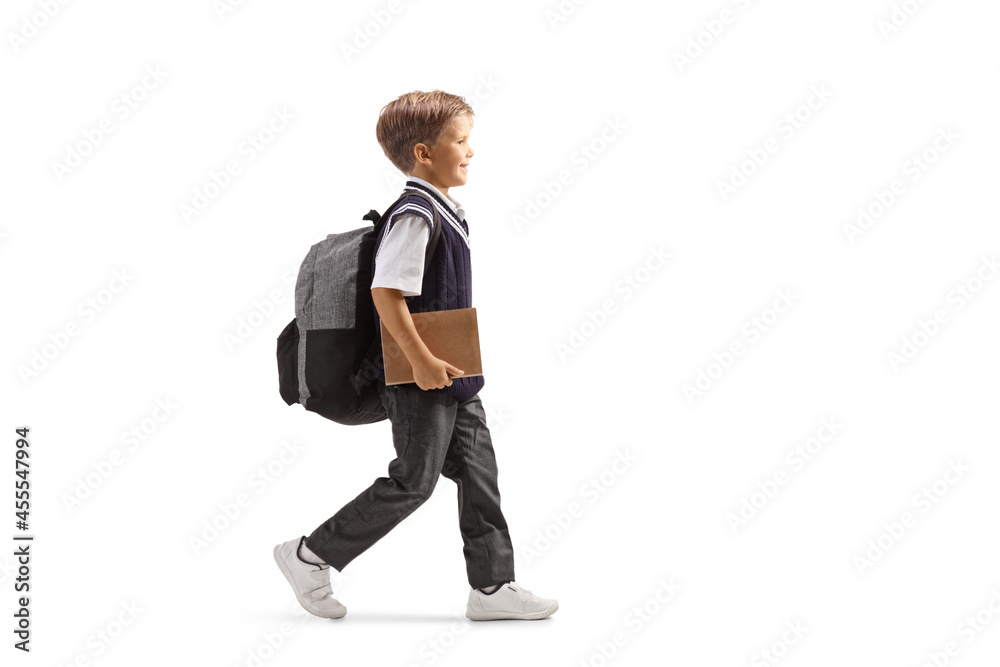 Full length profile shot of a little schoolboy in a uniform carrying a backpack and a book and walking Full length profile shot of a little schoolboy in a uniform carrying a backpack and a book and walking