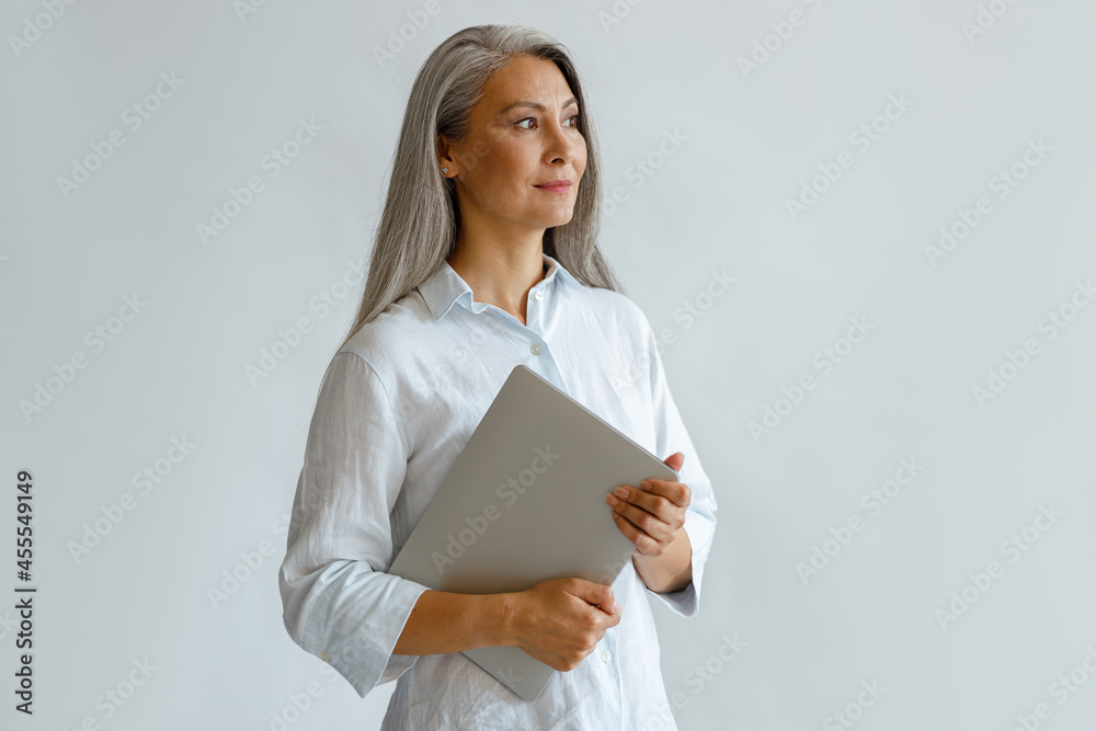 Positive mature Asian businesswoman wearing white blouse holds contemporary laptop standing on light grey background in studio Positive mature Asian businesswoman wearing white blouse holds contemporary laptop standing on light grey background in studio