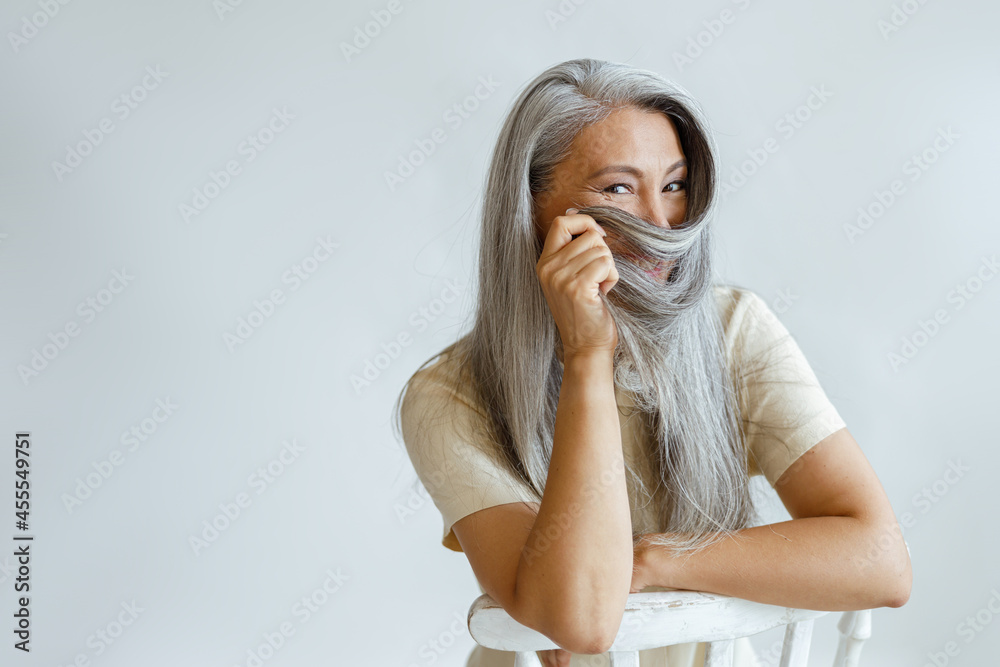 Positive middle aged Asian lady uses hoary hair as niqab sitting on chair on light background in studio, space for text. Mature beauty lifestyle Positive middle aged Asian lady uses hoary hair as niqab sitting on chair on light background in studio, space for text. Mature beauty lifestyle