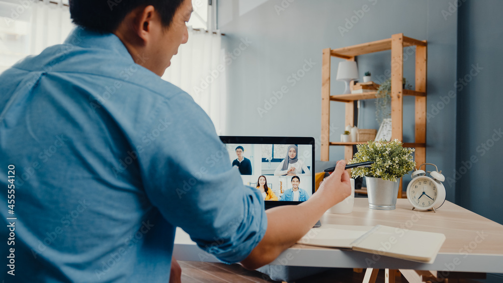 Fototapeta premium Young Asia businessman using laptop talk to colleagues about plan in video call meeting while work from home at living room. Self-isolation, social distancing, quarantine for corona virus prevention.