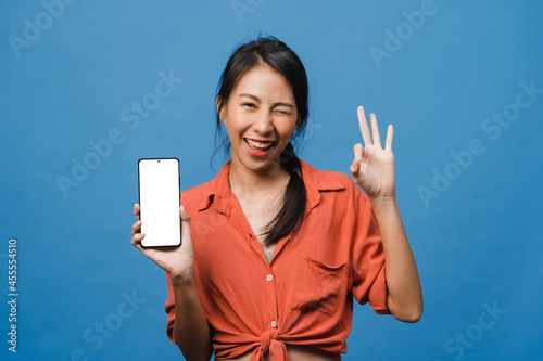 Young Asia lady show empty smartphone screen with positive expression, smiles broadly, dressed in casual clothing feeling happiness on blue background. Mobile phone with white screen in female hand. © tirachard