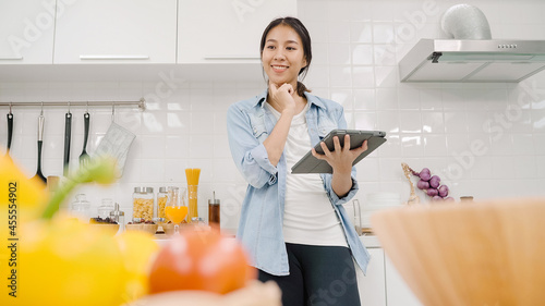 Happy Asian woman using tablet for looking recipe while making food in the kitchen, female use organic vegetable for healthy food at home. Lifestyle women making food concept.