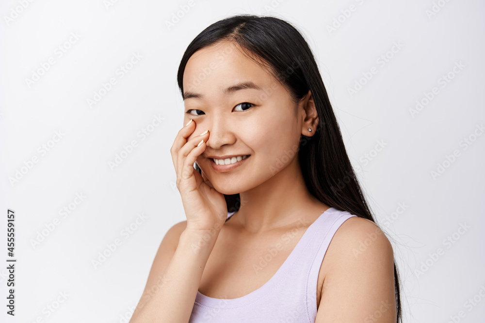 Attractive young asian woman with glowing healthy skin, smiling and touching face with fingertips, standing over white background Attractive young asian woman with glowing healthy skin, smiling and touching face with fingertips, standing over white background