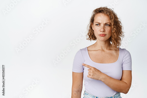 Suspicious young woman frowning, pointing finger and looking left with doubt, standing in t-shirt against white background