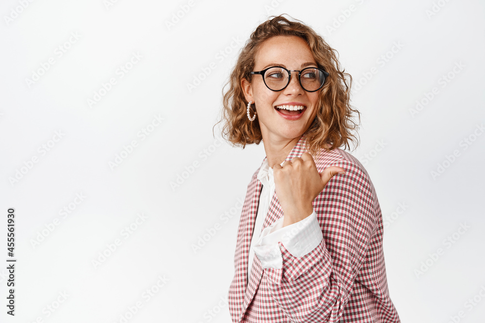 Smiling blond female entrepreneur, office manager in glasses and suit, pointing and looking right, behind her shoulder at copy space, white background